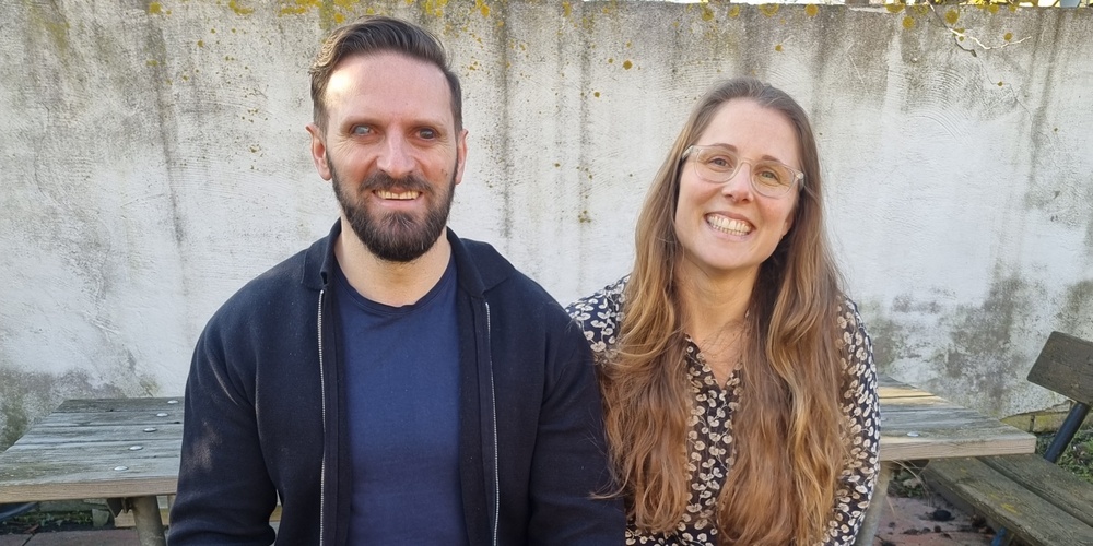 Två personer sitter på en bänk utomhus med en svart ledarhund mellan sig.