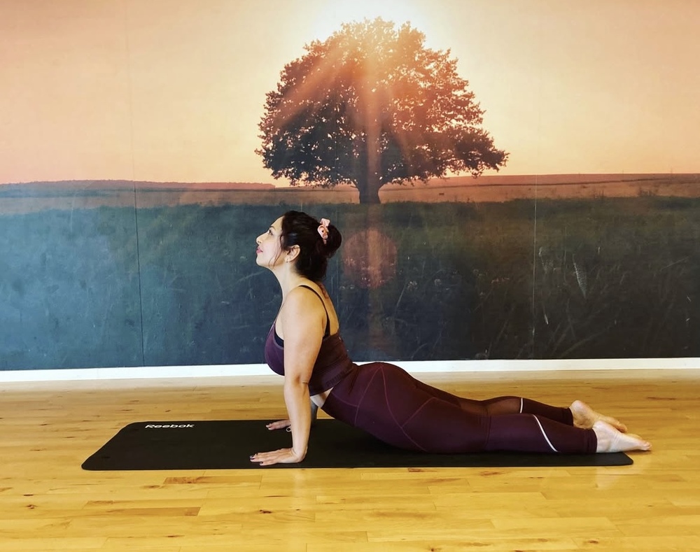 Kvinna gör en yogaposition på en matta i ett rum med trägolv och en väggmålning av ett träd i solnedgång.