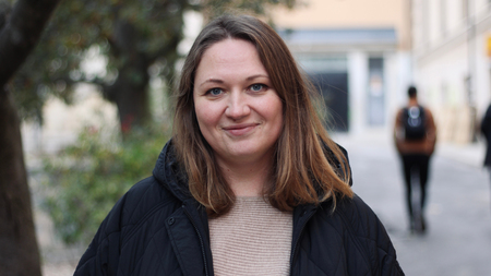 Kvinna med axellångt hår och svart jacka står utomhus på en gata, ler mot kameran.