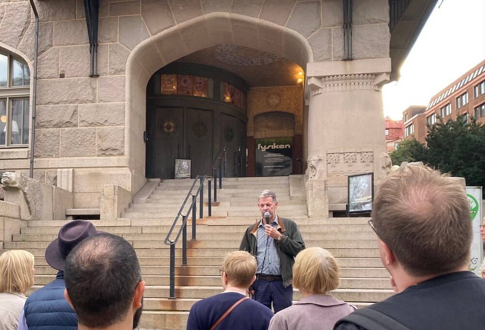 En man talar i mikrofon inför en liten publik utanför en byggnad med stentrappa och stora dörrar.