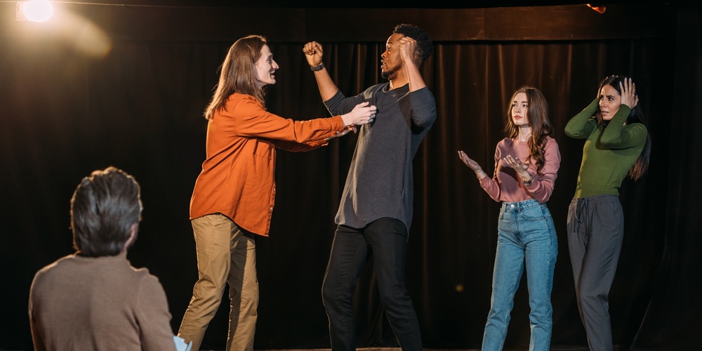 Fyra personer står på en scen och spelar teater, medan en person sitter och tittar på dem.