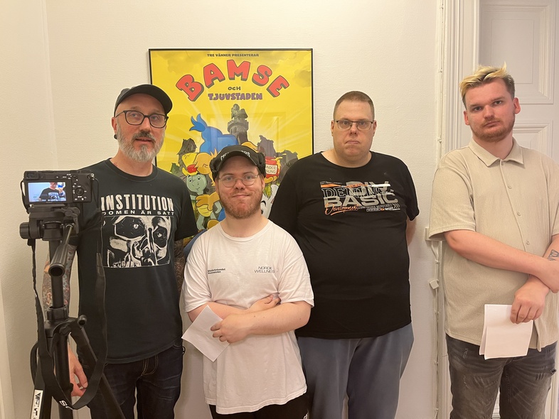 Fyra personer står framför en affisch med texten 'Bamse och tjuvstaden' i ett rum, en kamera på stativ syns till vänster.