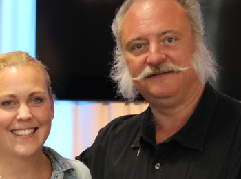 Två personer står tillsammans i ett rum med ljusa gardiner och en stor TV-skärm i bakgrunden.