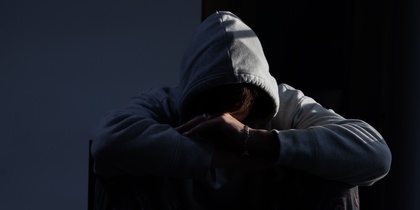 Person i grå hoodie sitter med huvudet nedböjt och armarna korsade, i ett mörkt rum.