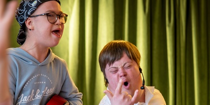 Två personer, en med gitarr och en som gör rocktecken, står framför gröna gardiner och ser glada ut.