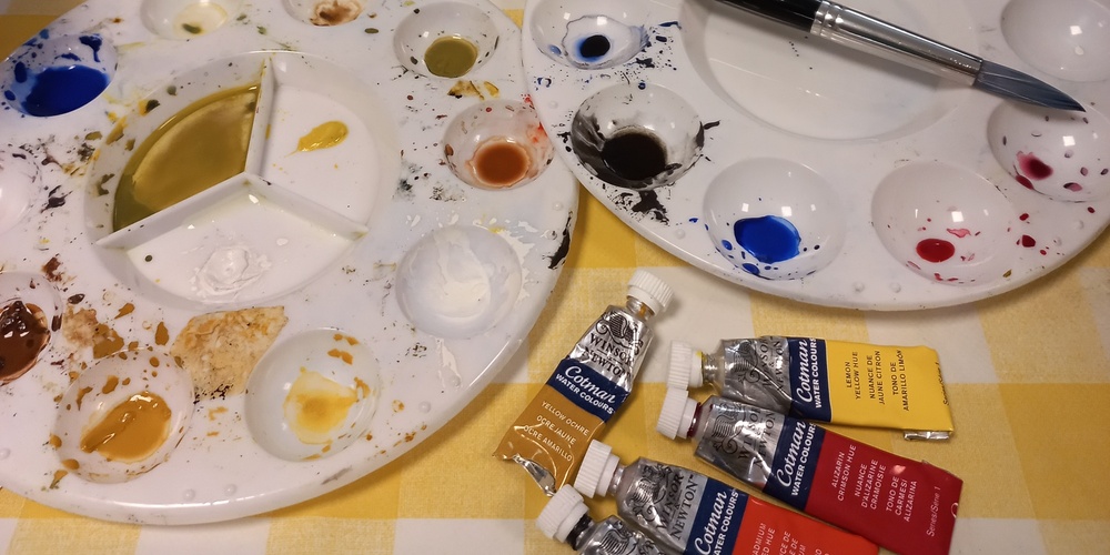 Två målarpaletter med färg, pensel, tuber med akrylfärg och två målningar på ett bord med gulrutig duk.