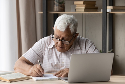 Äldre man med glasögon skriver i anteckningsbok vid laptop, omgiven av böcker på ett skrivbord.