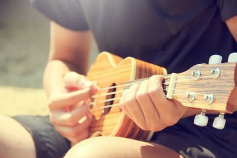 En person spelar ukulele utomhus i solljus, fokus på händer och instrument.