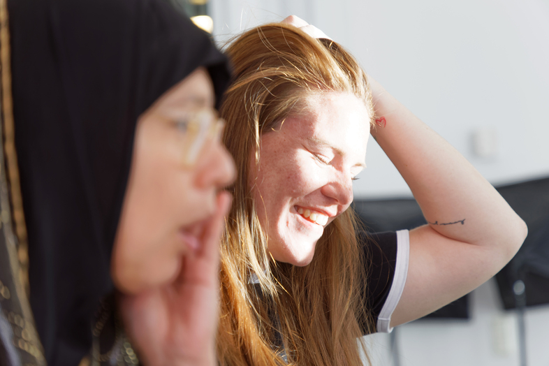Två kvinnor inomhus, en med hijab i förgrunden och en leende kvinna med långt hår i bakgrunden.