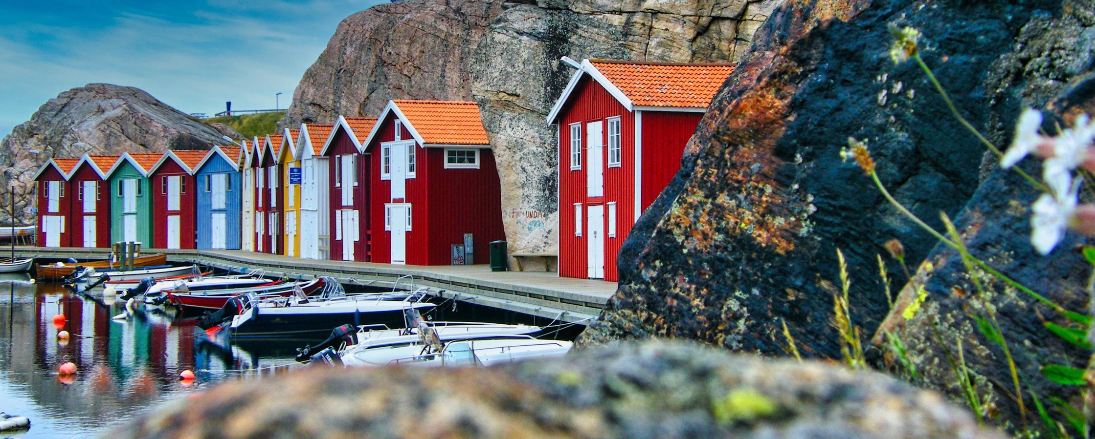 Färgglada sjöbodar vid vattnet med klippor i bakgrunden och båtar i förgrunden.