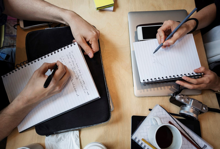 Två personer skriver i anteckningsböcker vid ett bord med kaffe, kamera och laptop.