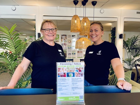 Två leende kvinnor står bakom en disk i en ljus och grönskande inomhusmiljö.