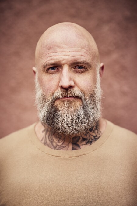 Skäggig, skallig man med tatueringar på halsen, beige tröja och neutral bakgrund.