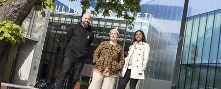 Tre personer står utomhus framför en modern glasbyggnad med träd och cyklar i närheten.