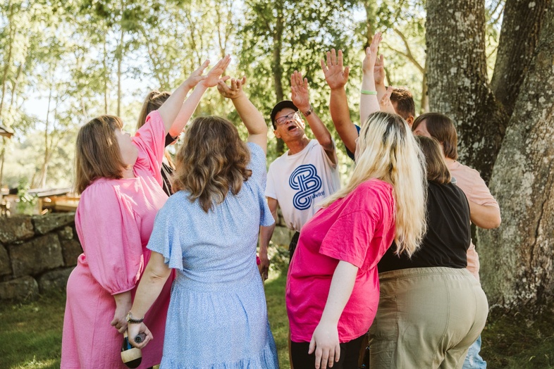 En grupp människor står i en ring utomhus och sträcker upp händerna mot mitten under ett träd.