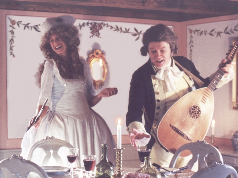 Två personer i 1700-talskläder vid ett dukat bord, en spelar luta och den andra sjunger eller skrattar.