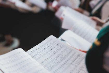 Närbild på ett notblad i en kör, med suddiga personer som håller sånghäften i bakgrunden.