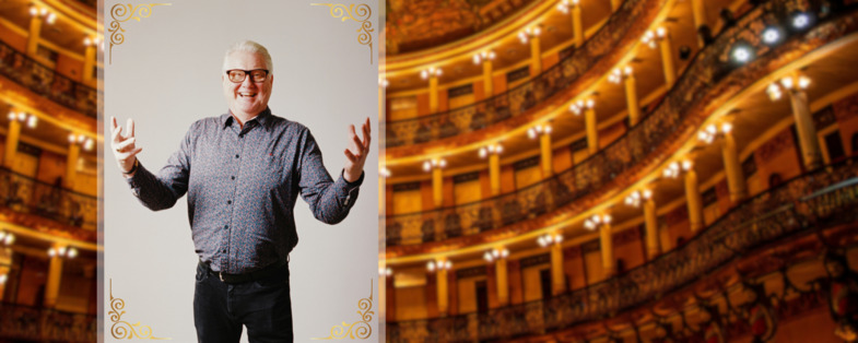 En leende man med glasögon står med utsträckta armar framför en bakgrund av en upplyst teatersalong.