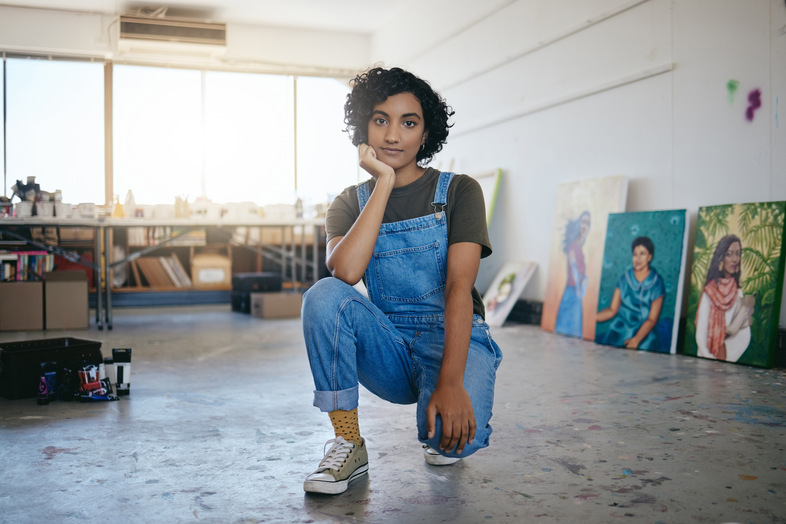 Ung kvinna i blå snickarbyxor sitter på huk i en konststudio med målningar i bakgrunden.