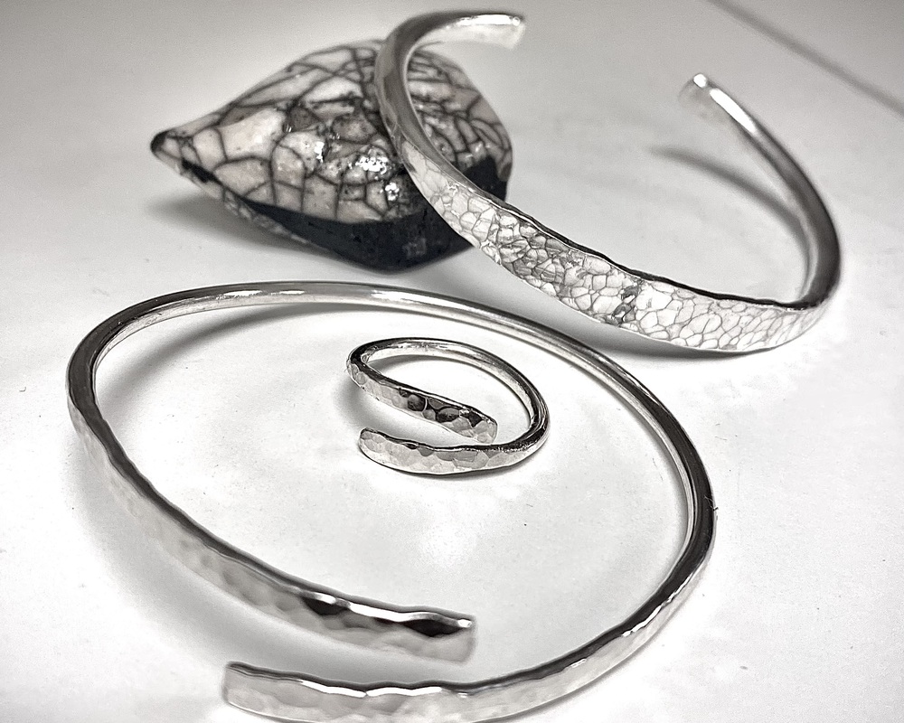Tre silversmycken, två armband och en ring, ligger på ett vitt underlag med en svartvit sten i bakgrunden.