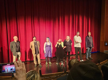 Sju personer står på en scen framför en röd ridå, med publik i förgrunden.