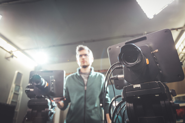 En person står suddigt i bakgrunden bakom två filmkameror i en studio med stark belysning.