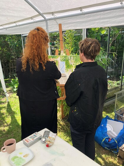 Två personer står under ett tält och målar vid ett staffli i en trädgård.