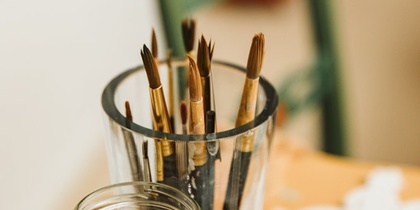 Flera penslar står i ett glas på ett bord, med suddig bakgrund och konstmaterial runtomkring.