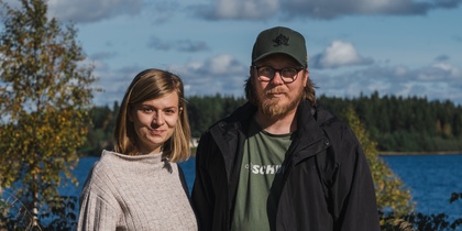 En kvinna och en man står utomhus vid en sjö med skog i bakgrunden under en solig dag.