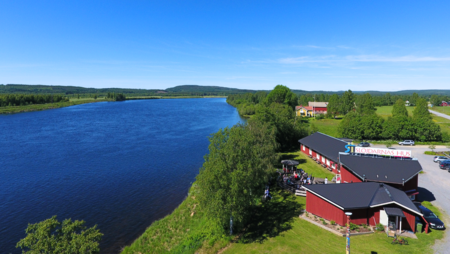 Landskapsvy med en bred flod, gröna ängar och röda byggnader under en klarblå himmel.