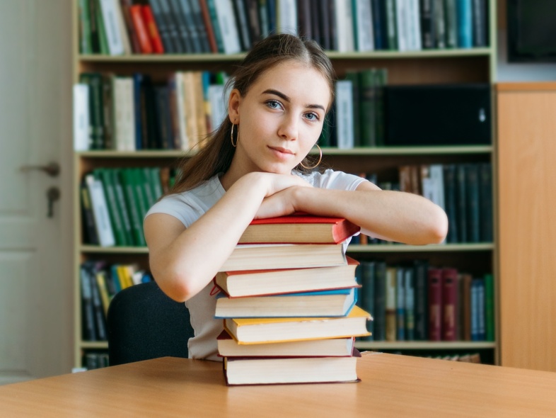 En ung kvinna sitter vid ett bord i ett bibliotek med en hög böcker framför sig.