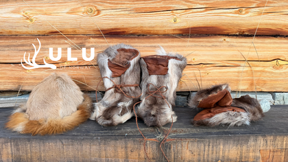 Traditionella samiska kläder av renskinn, inklusive mössa, skor och vantar, framför en timmervägg.