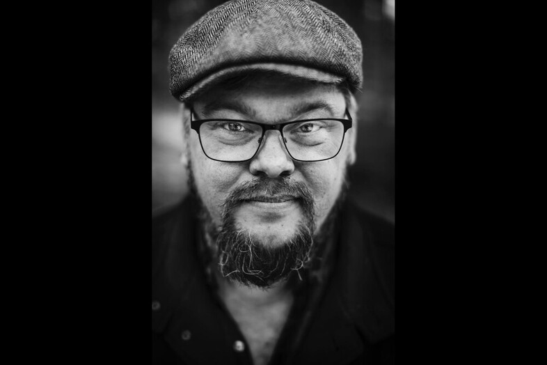 Porträtt av en man med glasögon, skägg och keps i svartvitt.