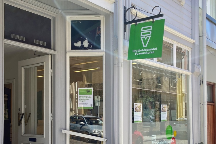 Entré till en glassbutik med grön skylt på en solig gata, bänk utanför och stora fönster.