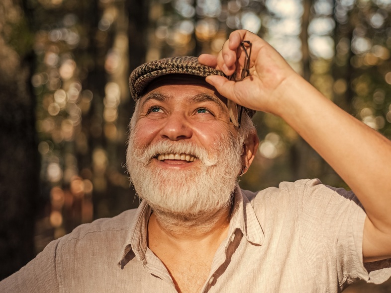 Leende äldre man med vitt skägg och keps står utomhus i soligt skogsparti.