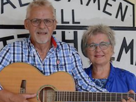 En äldre man med gitarr och en äldre kvinna står tillsammans framför en skylt, båda ler mot kameran.
