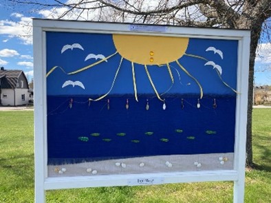 En anslagstavla utomhus visar en konstnärlig bild av sol, hav, fåglar och fiskar gjorda av olika material.