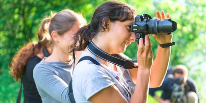 Två kvinnor fotograferar utomhus i grönskande miljö, med en tredje person i bakgrunden.