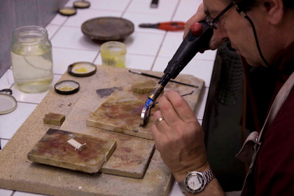 En person arbetar med en liten gasbrännare vid ett arbetsbord, troligen inom smyckestillverkning eller metallarbete.