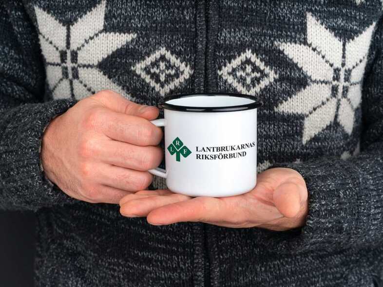 En person i en grå kofta med vitt mönster håller en emaljerad kopp med LRFs logotyp framför kroppen med båda händerna.