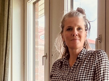 Person i svartvit mönstrad skjorta står vid ett fönster i ett ljust rum med gröna gardiner och papperspåsar.