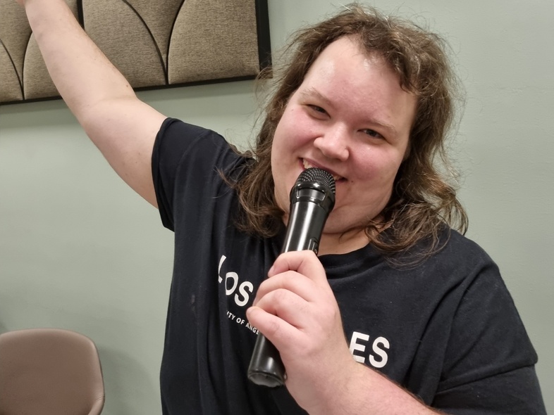 Person sjunger i mikrofon med ena armen uppsträckt, står i ett rum med ljusgrön vägg och ljudabsorbenter.