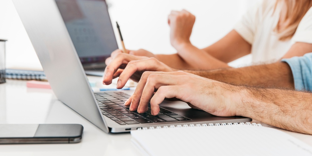 Två personer arbetar vid en bärbar dator med anteckningsblock och mobiltelefon på ett vitt skrivbord.