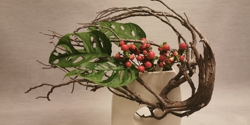 En modern blomsterarrangemang i vit vas med gröna blad, röda bär och böjda grenar mot ljus bakgrund.