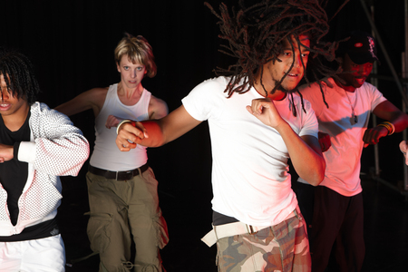 Fyra personer dansar energiskt på en scen, klädda i vita och ljusa kläder mot en mörk bakgrund.