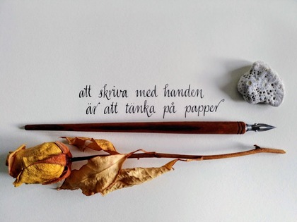 En handskriven svensk text på papper med en gammal bläckpenna, en torkad ros och en sten bredvid.