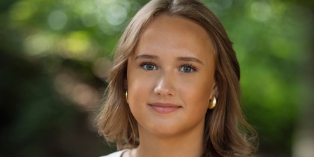 Person med axellångt brunt hår och vit topp står utomhus med grönskande bakgrund.