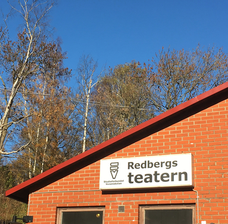 Röd tegelbyggnad med skylt som det står 'Redbergs teatern' på, träd och blå himmel i bakgrunden.