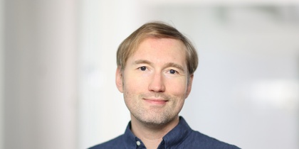 Man med ljust hår och blå skjorta står framför en ljus, suddig bakgrund.