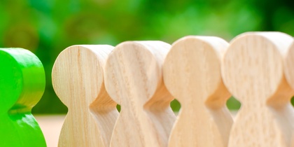 En grön träfigur står mellan två rader av ljusa träfigurer på ett bord med grön bakgrund.